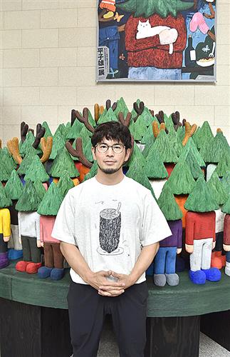 頭が木で身体が人間のキャラクターの作品の前に立つ平子雄一さん